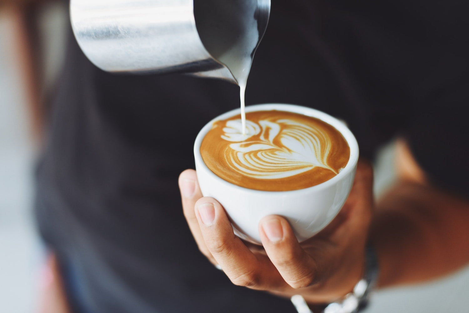 A person pouring milk into a coffee drink, creating a pattern on the surface, indicative of a barista making a latte or cappuccino.